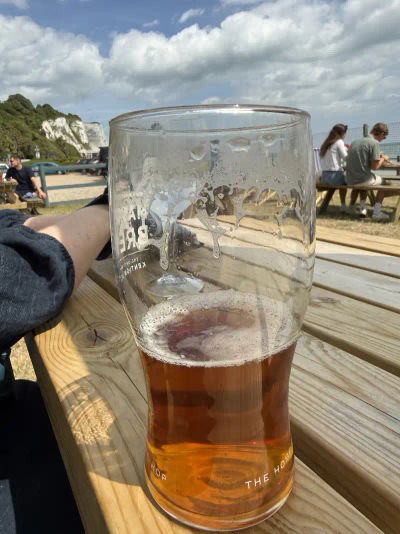 Pint of Master Brew beer on a wooden slatted outdoor beer garden table with the White Cliffs and English Channel in the background and puffy white clouds on a bright blue sky
